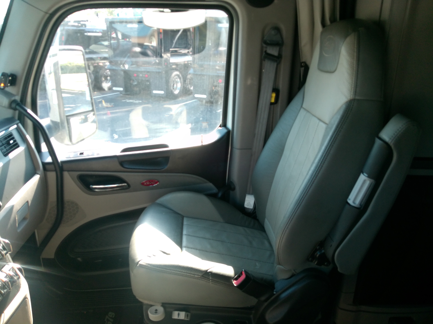 Truck cabin interior of a Peterbilt with a gray driver seat, door panel, and window view showing a reflection of other trucks.
