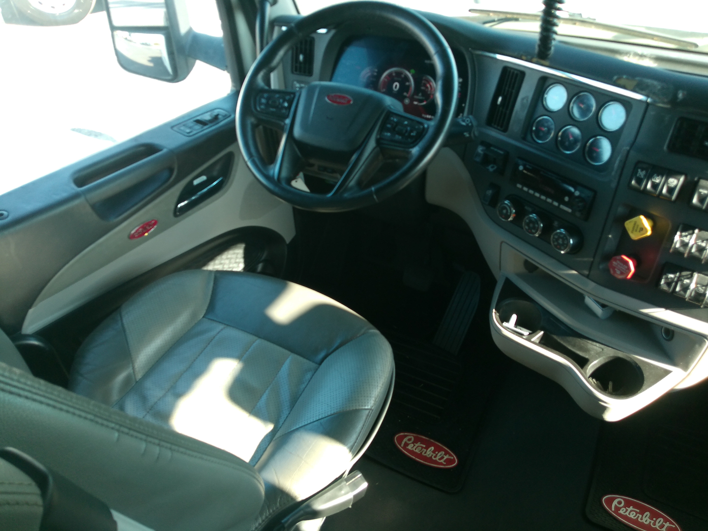 Peterbilt truck interior with steering wheel, dashboard gauges, and center stack visible; floor mat shows Peterbilt logo.