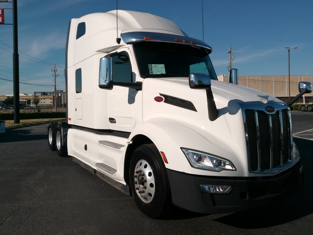 Peterbilt 579 conventional sleeper truck in white with chrome grille and dual mirrors, front right view. Visible text on side window sticker