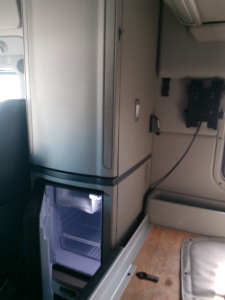Truck interior showing a vertical refrigerator/freezer unit and a small open compartment; cab area with wiring panel and seating visible.