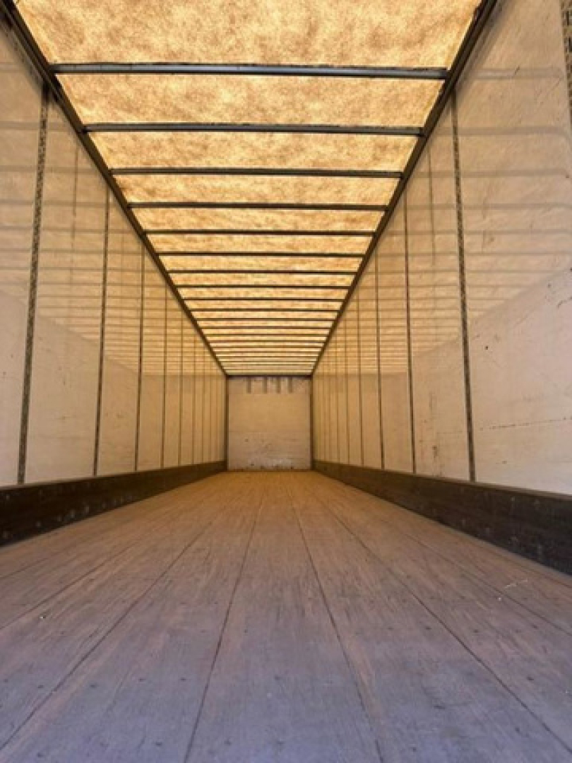 Inside view of a long empty trailer with wooden floor and white interior walls, leading to a back door; no text visible.