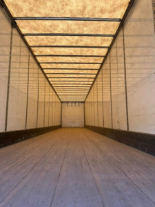 Inside view of a long empty trailer with wooden floor and white interior walls, leading to a back door; no text visible.