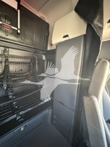Truck cab interior showing storage and seating area from a Freightliner Cascadia 126 sleeper, with panels, shelves, and a light-colored seat in view.