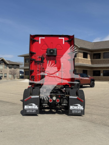 Rear view of a red Freightliner truck tractor with dual rear fenders and license area, viewed in a parking lot near a building.