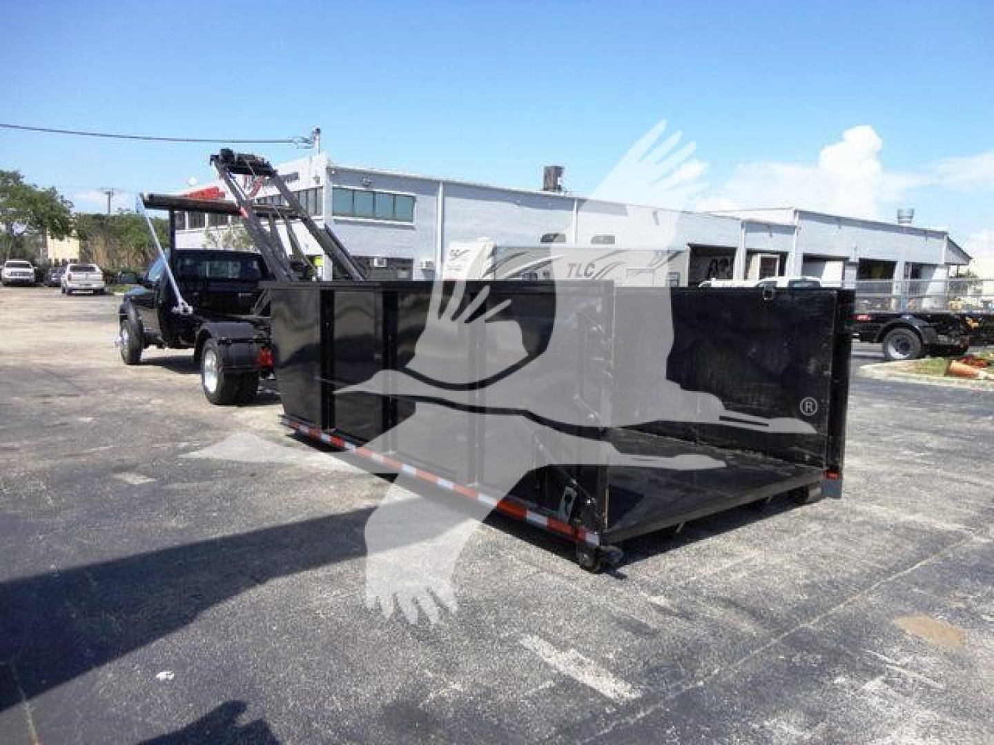 Ram 5500 dump truck in a lot with a large black roll-off dumpster attached, side view showing cargo bed and hoist mechanism.