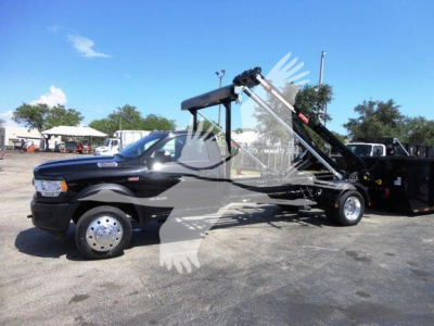 Ram 5500 HD tow truck with a roll-off hoist, black cab, chrome wheels, blue sky, industrial lot in background.