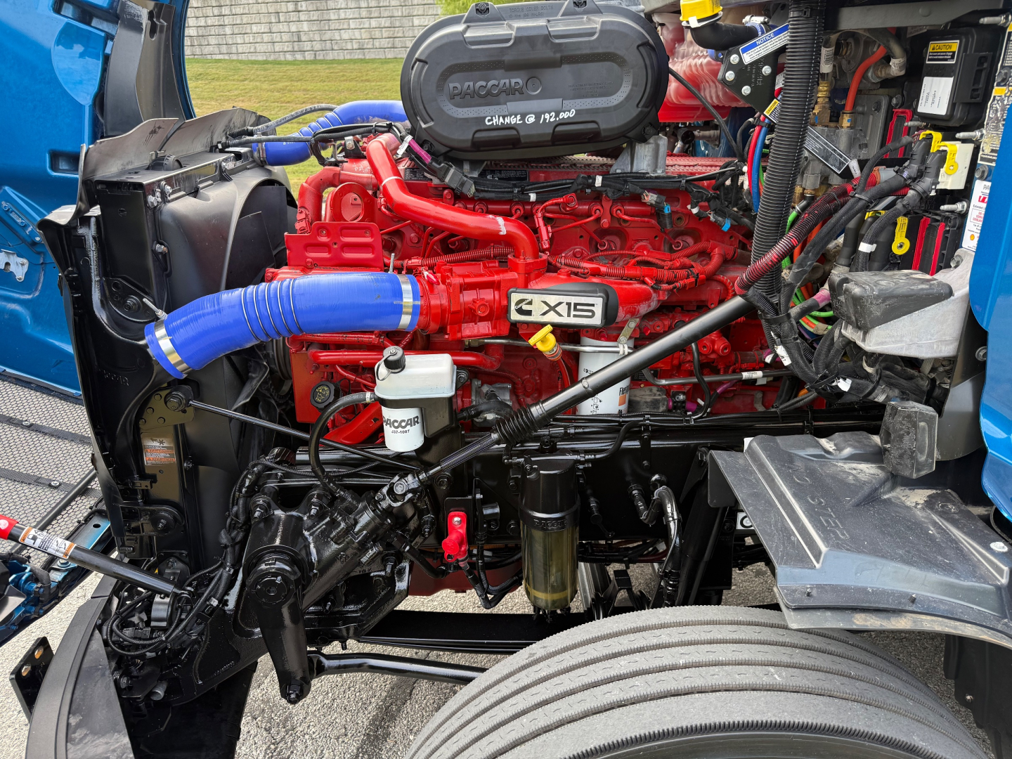 Peterbilt 579 engine bay showing red Cummins 12.0L engine with blue radiator hose, air intake, filters, and surrounding components.