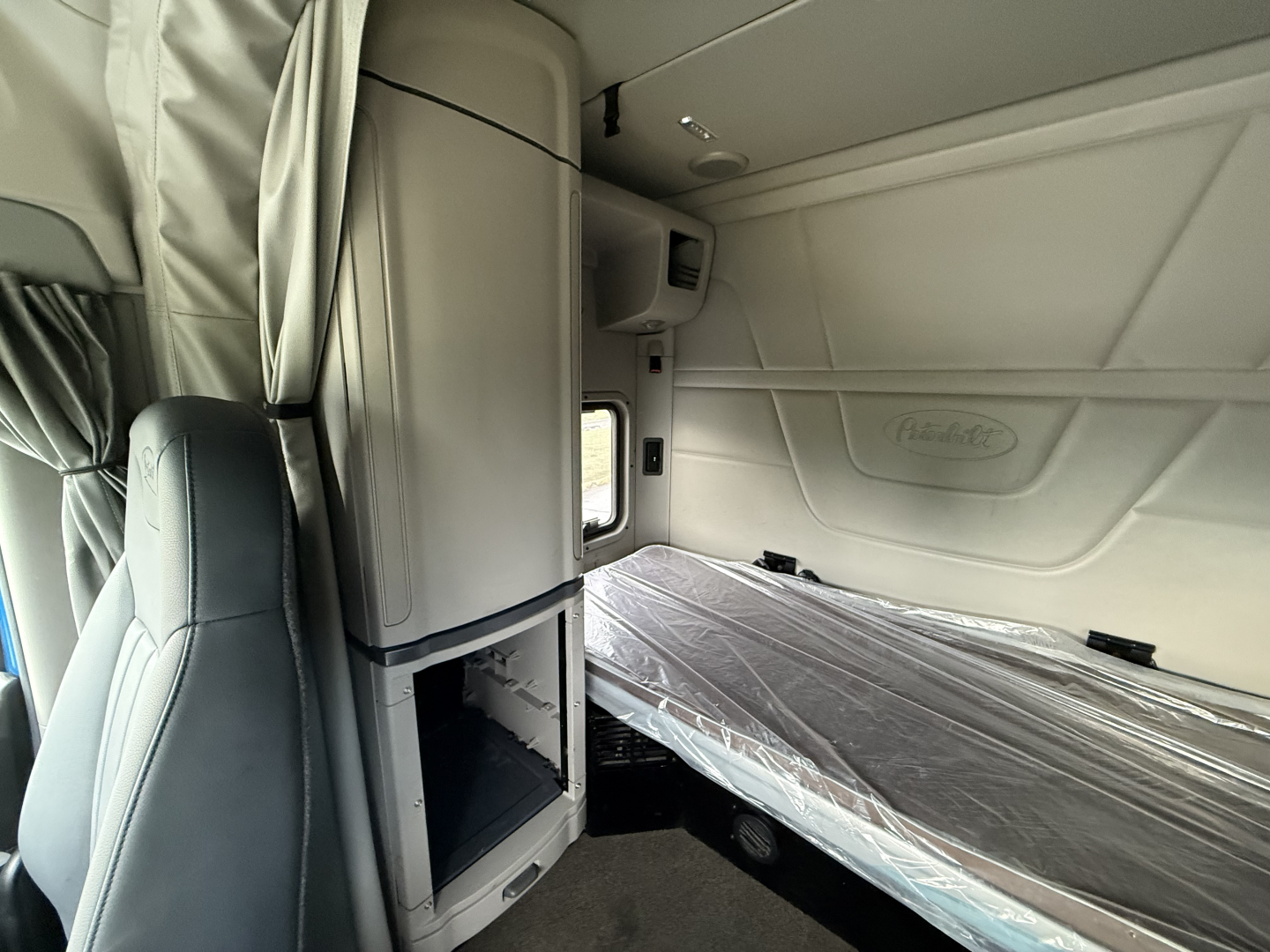 Peterbilt 579 sleeper cab interior showing side bunk bed with plastic cover, cabinet above, window and light; part of dash and seating visible.