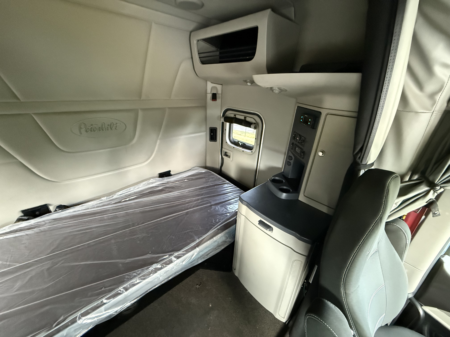 Peterbilt sleeper cab interior showing bunk bed with plastic cover and side console; window and control panels visible.