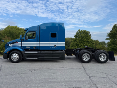 Side view of a blue 2025 Peterbilt 579 conventional sleeper truck, with white stripes along cab, shown on a paved lot.