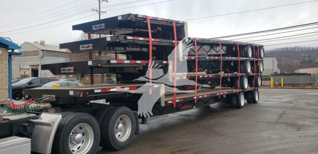 Heavy-duty trailer with multiple leaf-sprung decks and stacked steel frames loaded on a tri-axle trailer showing spare tires and red straps.