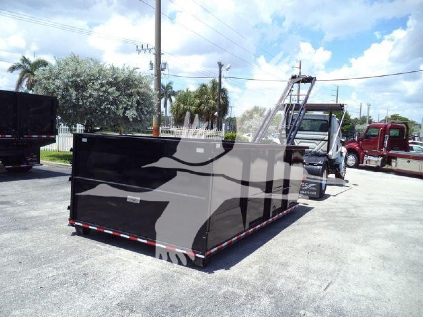 Truck with large black roll-off dumpster in a lot, semi-truck cab in background, blue sky with clouds.