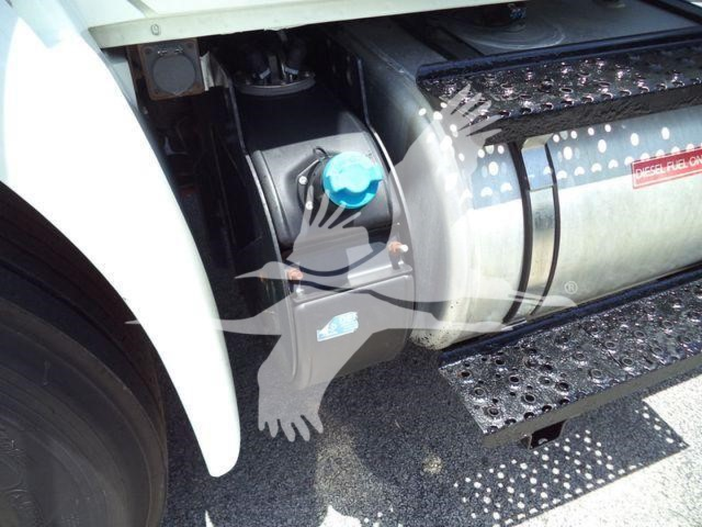 Truck engine bay showing blue cap on fluid reservoir and cylindrical component near front wheel area.