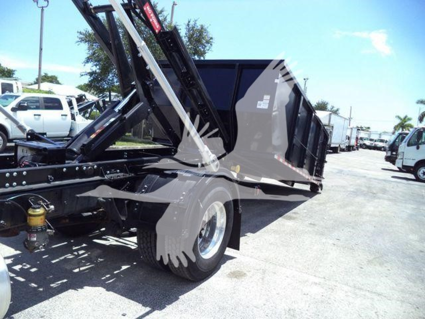 Truck chassis and dump bed of a 2020 International MV roll-off, side view with hydraulic hoist, yard with other trucks in background.