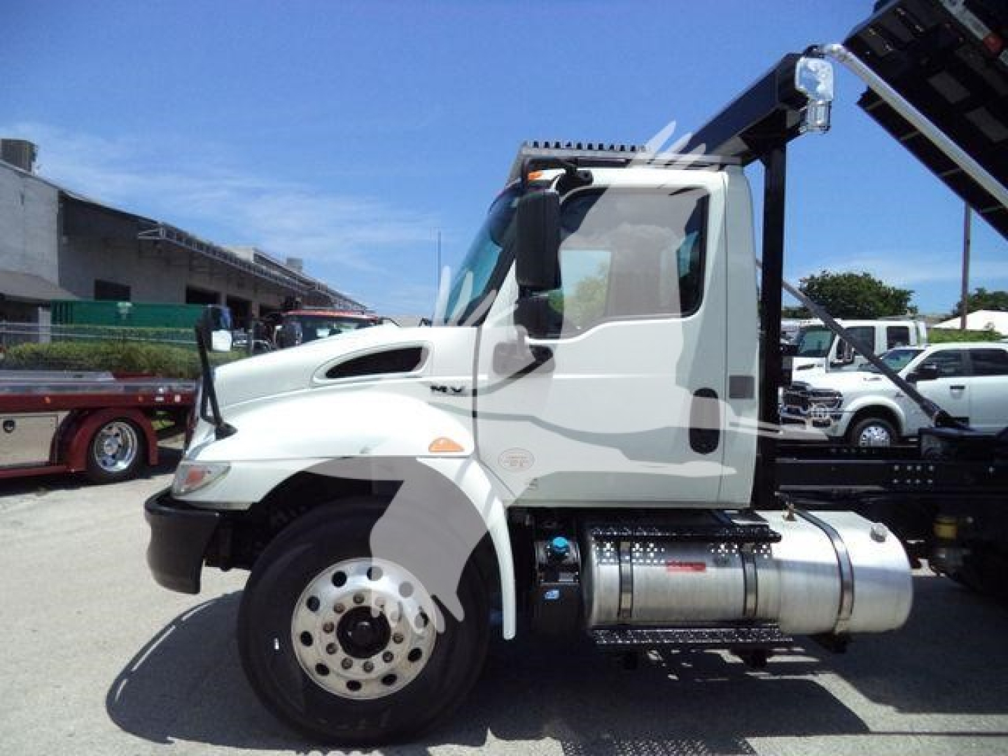 Truck side view of a 2020 International MV roll-off chassis showing cab, fuel tank, and wheel; clear sky and dealership lot in background.