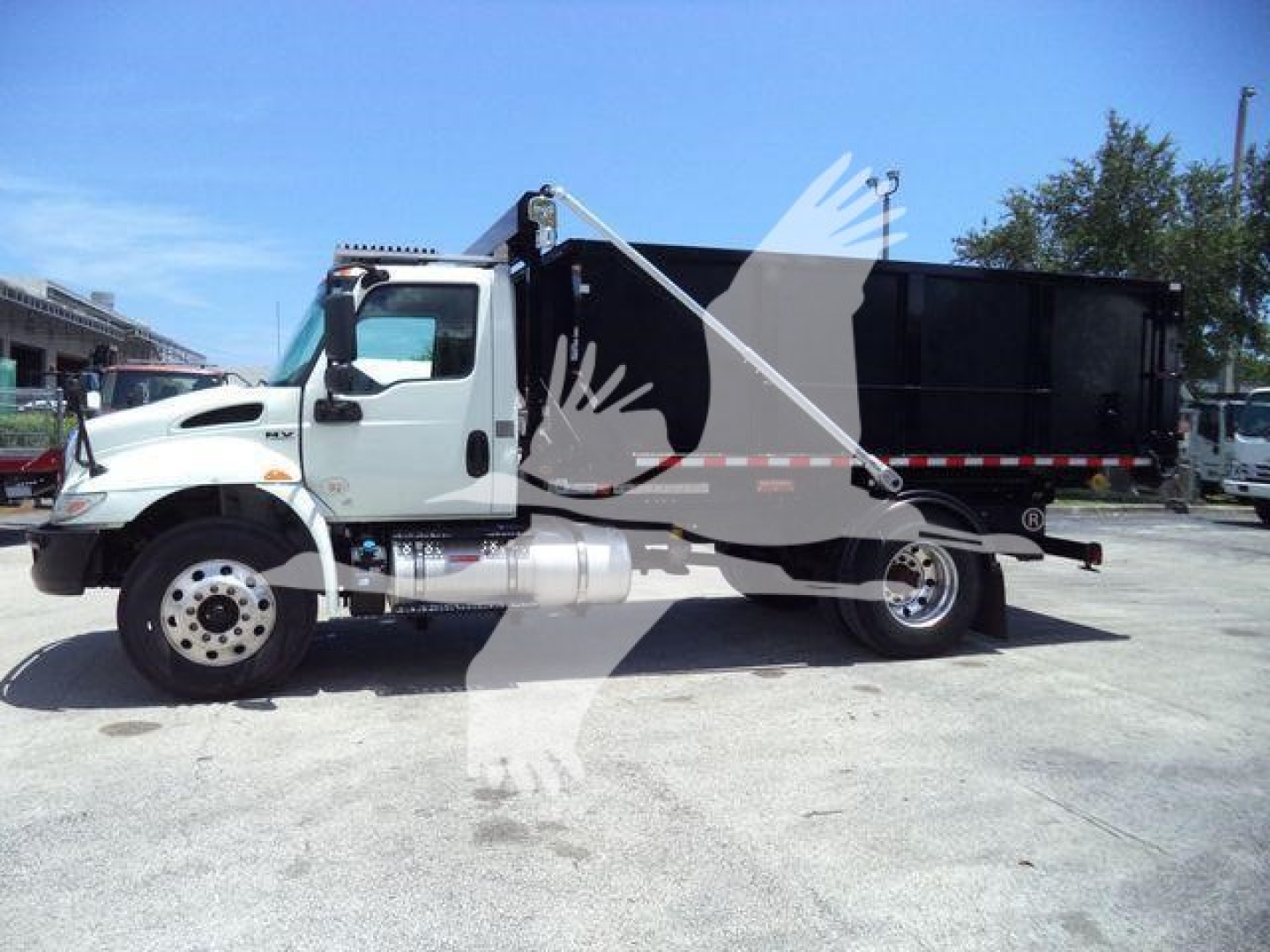 Truck side view showing a white International MV roll-off truck with black dump body and ladder, no visible branding text readable.