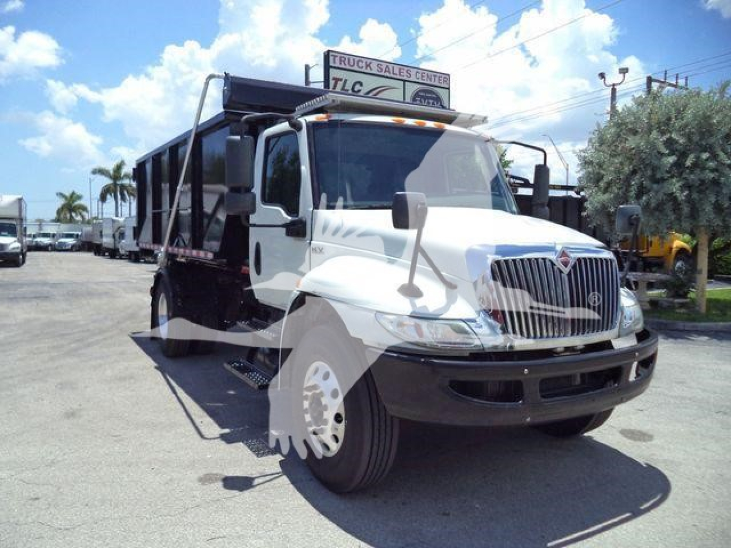 International MV roll-off truck in white and black with dump bed, front grille, large side mirrors, and visible front wheel; dealership lot in background.