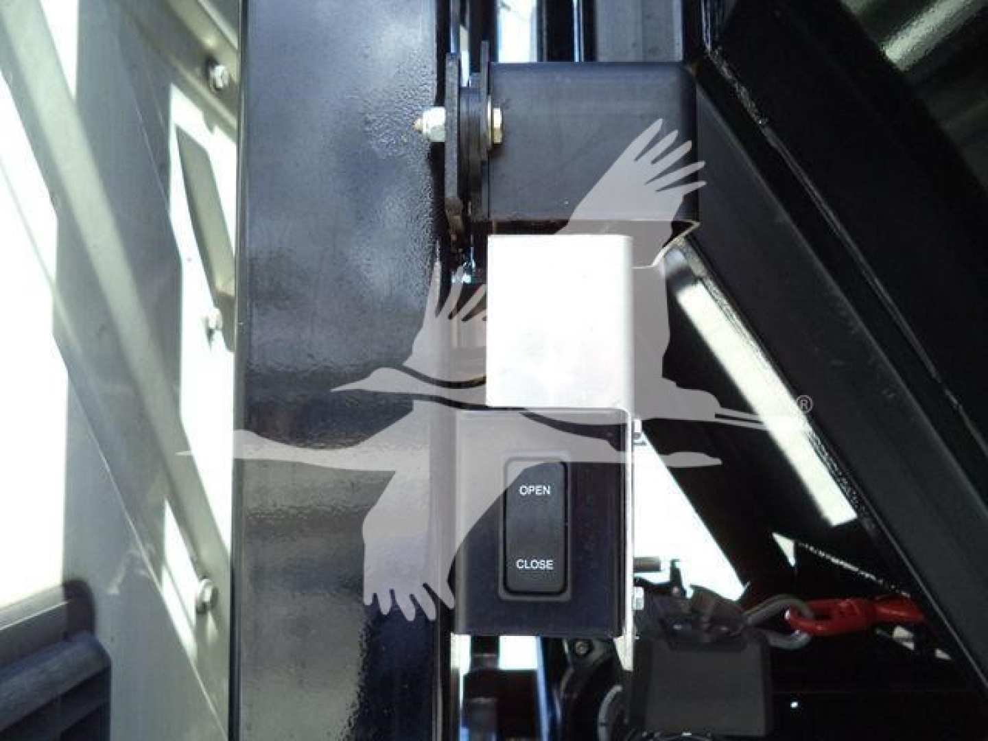 Close-up showing a switch labeled OPEN/CLOSE mounted on a metal frame with hydraulic lines and a lever in a rolloff truck. Partial view of machinery and a white square label.