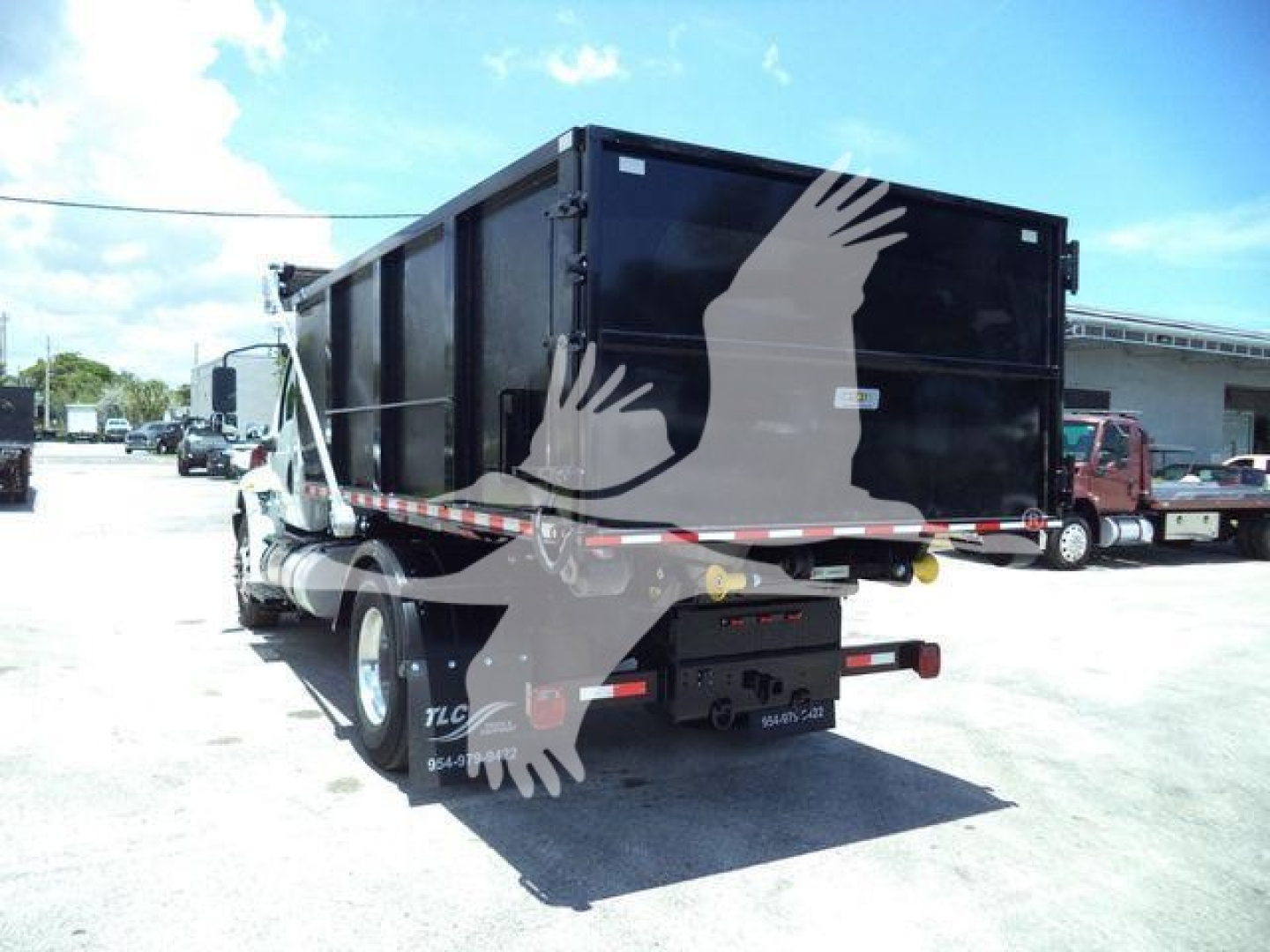 Back view of a black dump/roll-off truck trailer with double rear wheels, rear hitch, and diagonal reflective tape. Some text on mudflap and plate area partially visible.