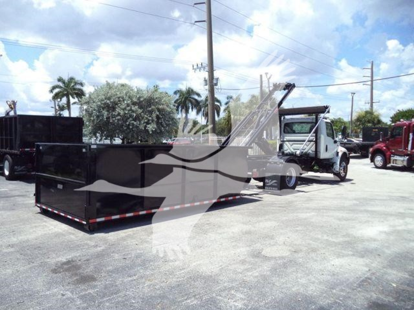 Truck with large black roll-off container and hydraulic arm in service bay showing MV series dump bed and dealership lot.