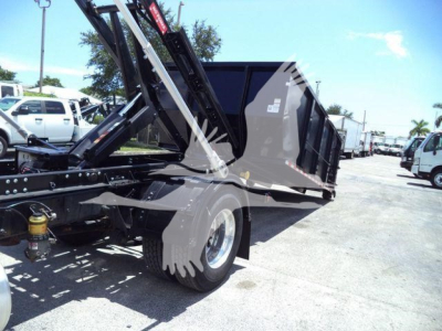 Truck chassis and dump bed of a 2020 International MV roll-off, side view with hydraulic hoist, yard with other trucks in background.