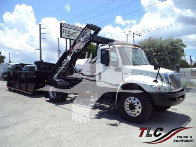 Truck cab with roll-off hoist in a used 2020 International MV, front and side view showing loading container and TLC Truck Sales sign in the background.