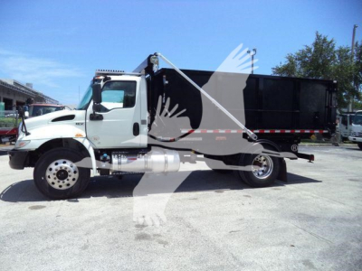 Truck side view showing a white International MV roll-off truck with black dump body and ladder, no visible branding text readable.