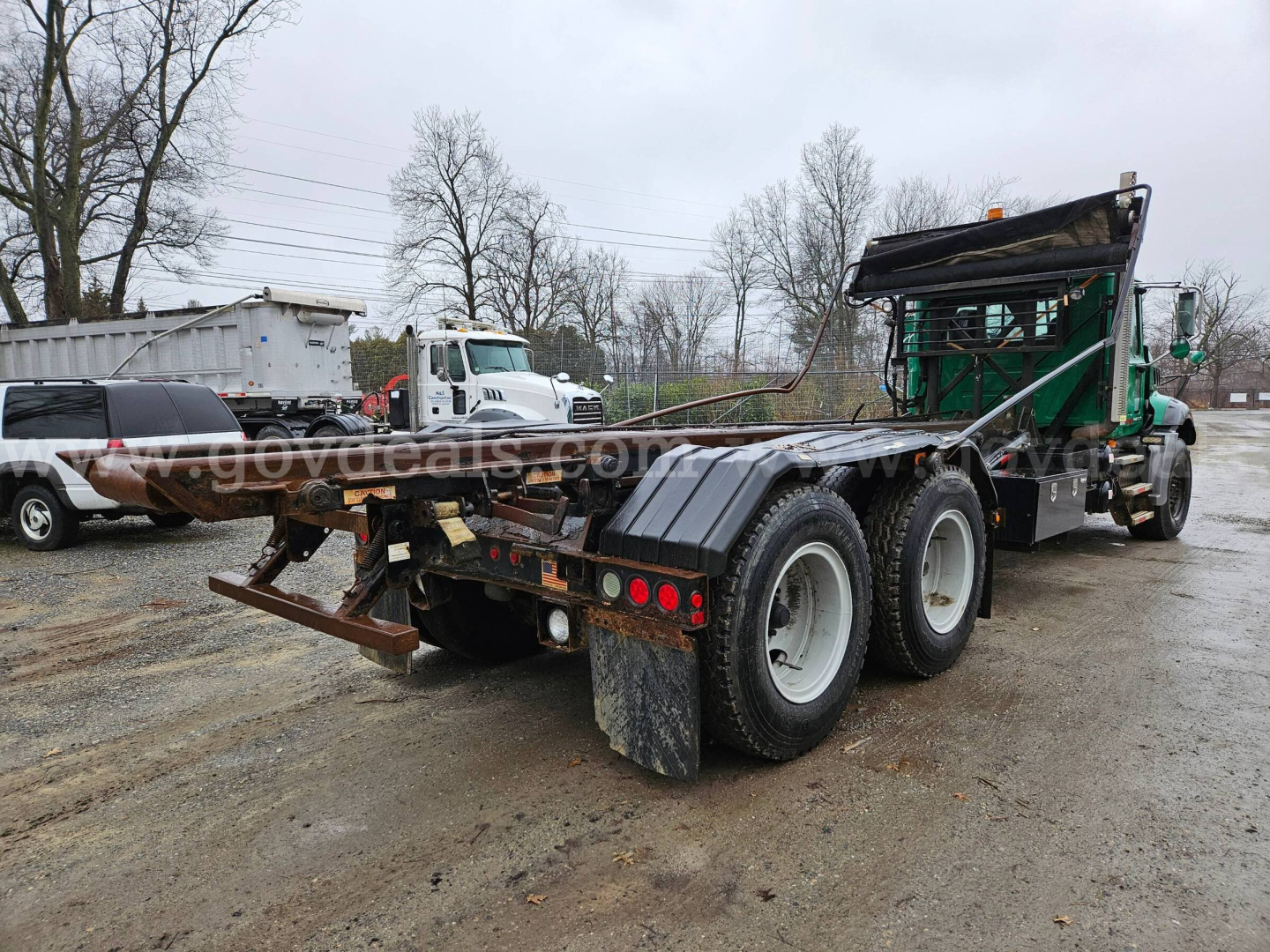 Mack GU813 rolloff truck chassis from rear-right view showing dual rear axles and flatbed bed, hydraulic system, and green cab in background.