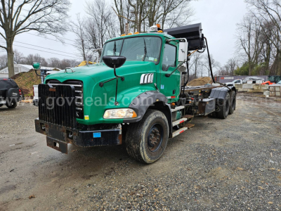 Green Mack GU813 roll-off truck with front bumper, black grille, dual rear axles, and a hydraulic dump bed in an outdoor lot.