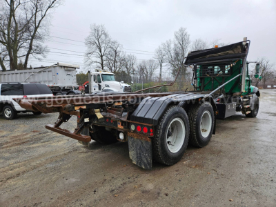 Mack GU813 rolloff truck chassis from rear-right view showing dual rear axles and flatbed bed, hydraulic system, and green cab in background.