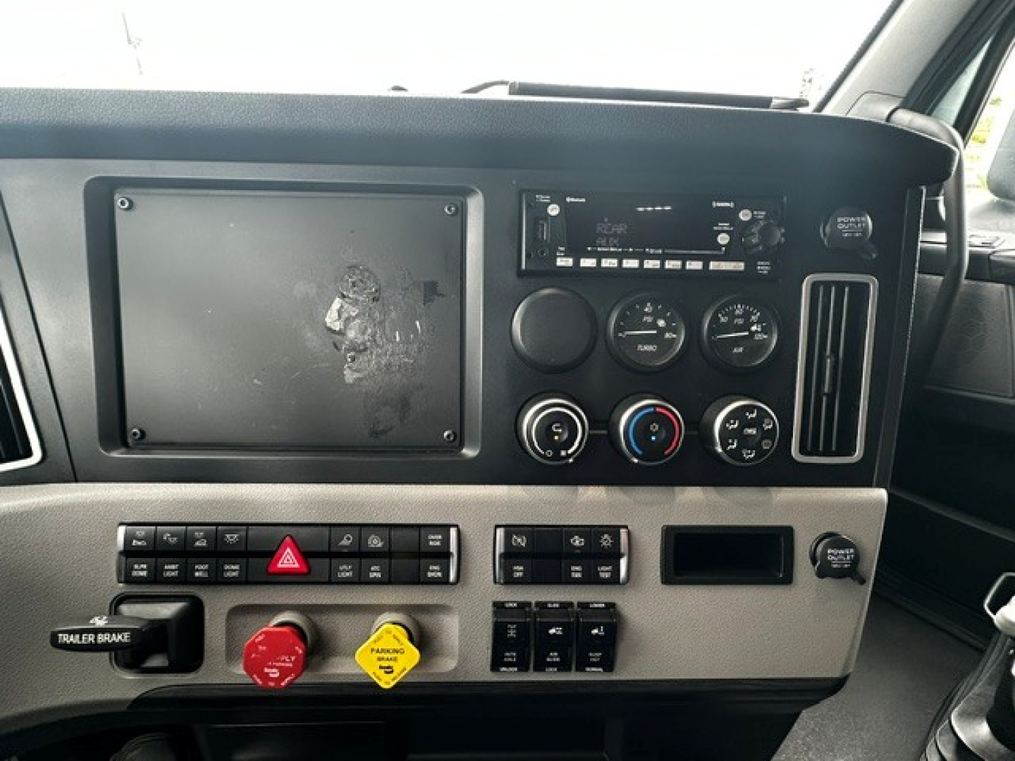 Dashboard and center console of a Freightliner Cascadia 126 showing a large blank display screen, climate control dials, radio, various switches, hazard button, trailer brake, and yellow warning knob.