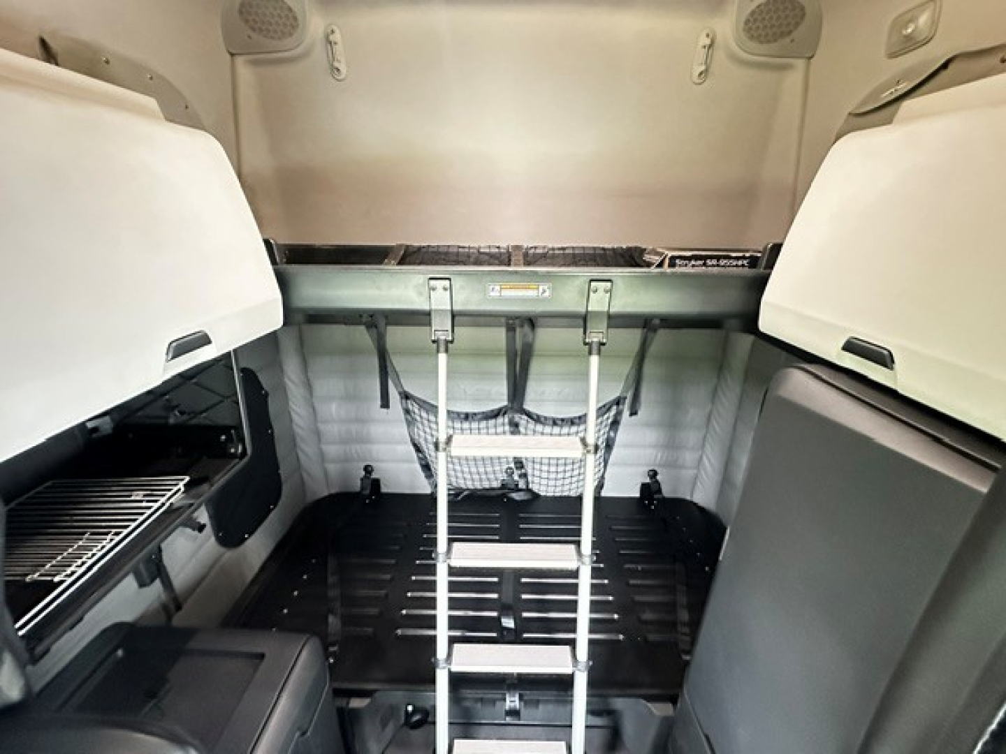 Freightliner Cascadia 126 sleeper cab cargo area with overhead storage and ladder in the back, interior view showing tie-down hooks and side compartments.