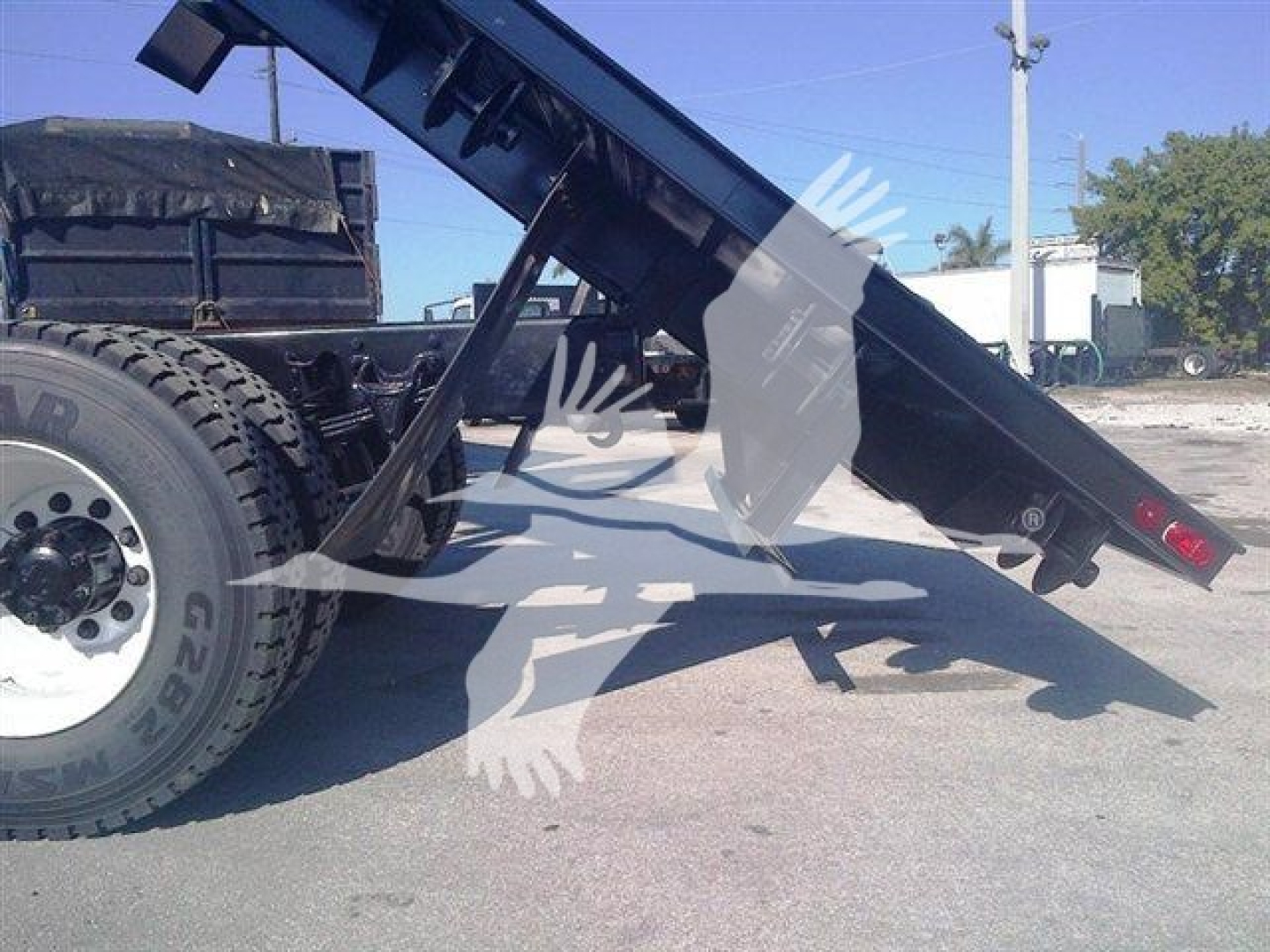 Flatbed trailer hitch and hydraulic tilt assembly attached to a truck chassis showing steel frame and tire with fender.