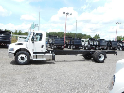 White 2027 Freightliner M2 106 cab and chassis showing side view with empty chassis, logo on door, parking lot with other trucks in background.