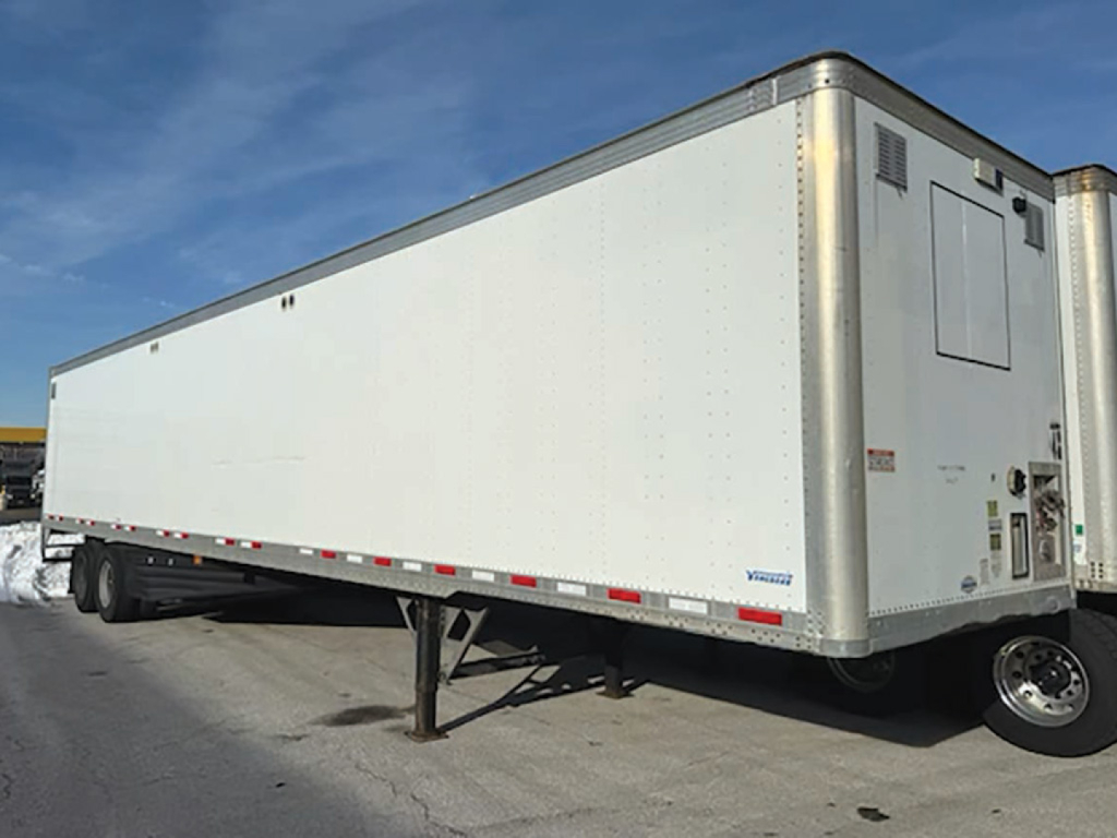 Trailer side view of a 2020 Vanguard aluminum box van with landing legs down and a front cab area at the right