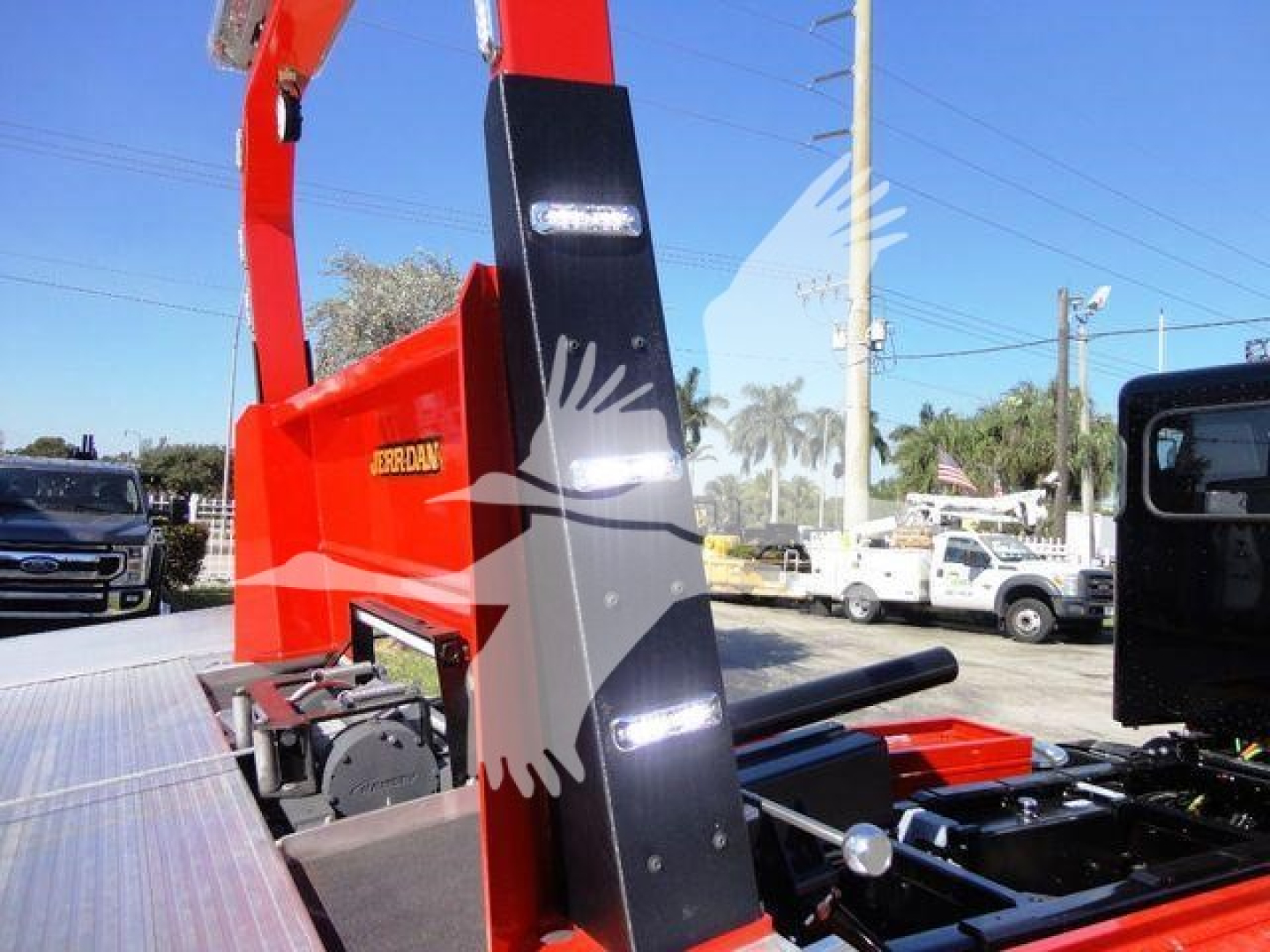 Peterbilt 337 rollback apparatus with orange-red hydraulic boom, side display panels, and warning lights; visible text includes 'JORDAN' on the side of the boom and surrounding equipment.
