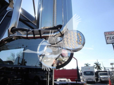 Peterbilt 337 truck cab mirror with round LED strobe/spotlight assembly showing wiring and chrome mounting, trucks in background.