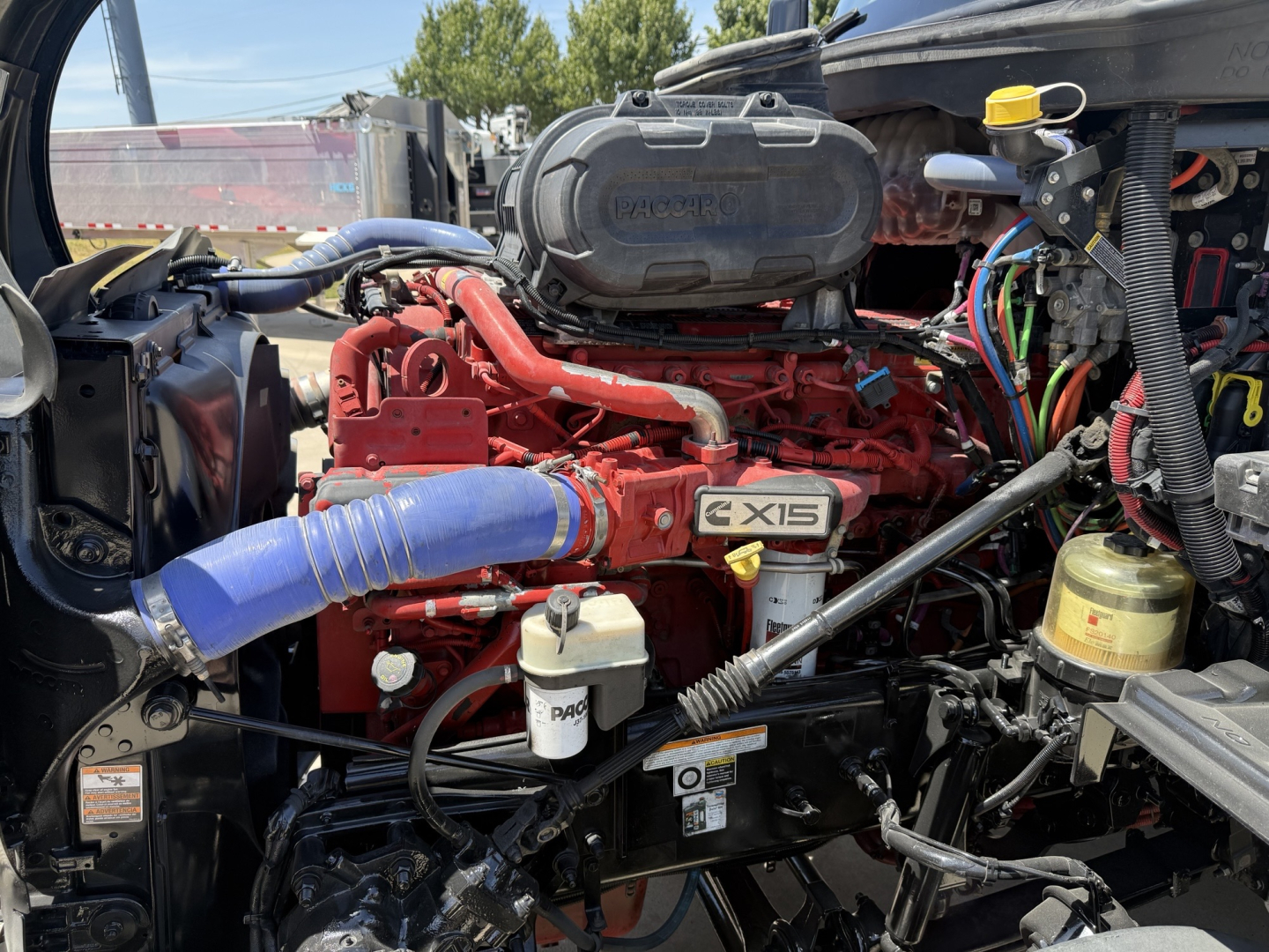 Peterbilt 579 engine bay showing Cummins X15 engine with blue intake hose and various hoses and reservoirs.