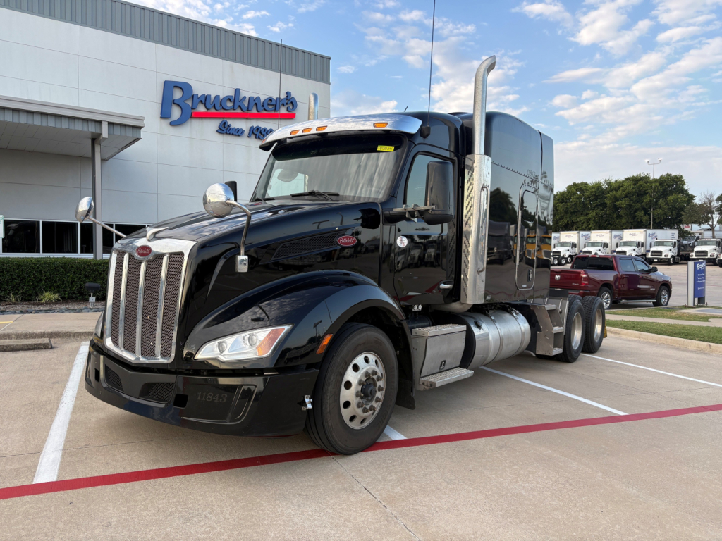 Peterbilt 579 conventional sleeper tractor in black, front three-quarter view, chrome grille, dual exhaust stacks, daytime running lights, dealership in background.
