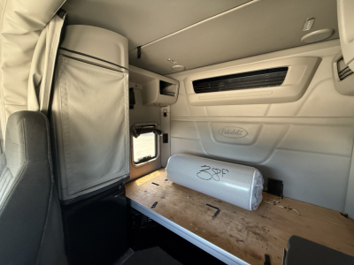 Peterbilt 579 interior sleeper cab with upper storage, window, and a rolled cylindrical bolster on a wooden bunk shelf.