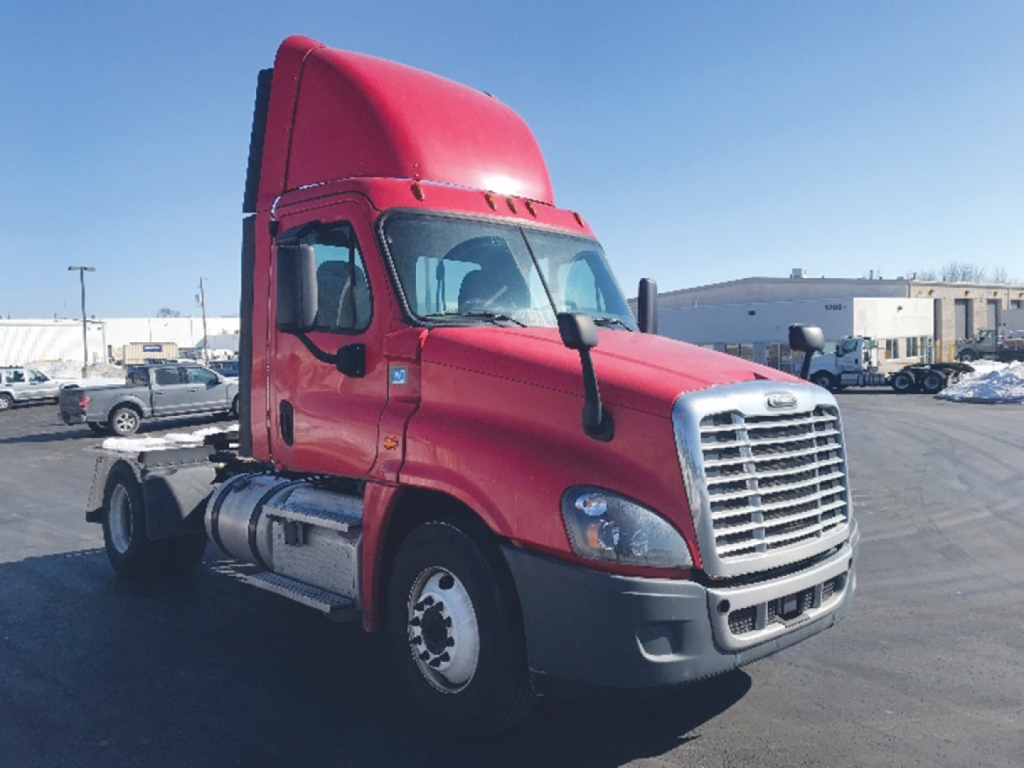 Truck cab showing long-nose Freightliner Cascadia125 with red exterior, side mirrors, chrome grille, and dual fuel tanks in a parking lot.