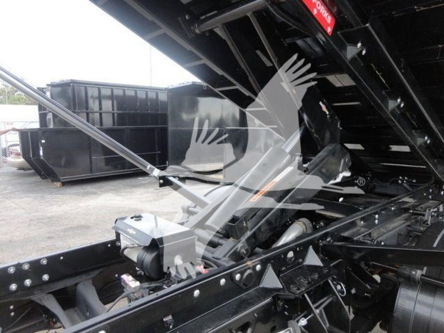 Ram 5500 SLT truck in a hooklift configuration showing chassis, hydraulic components, and transfer linkage with a large black dumpster in the background.