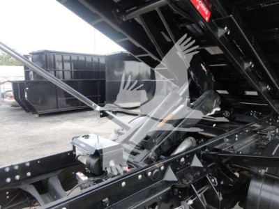 Ram 5500 SLT truck in a hooklift configuration showing chassis, hydraulic components, and transfer linkage with a large black dumpster in the background.