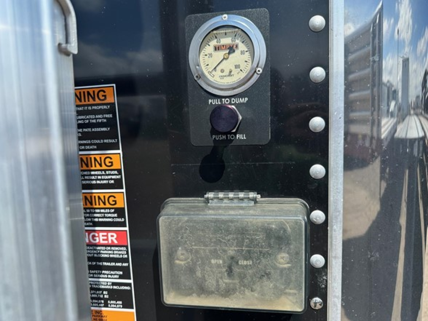 Hopper trailer control panel with a round gauge labeled 'PULL TO DUMP' and 'PUSH TO FILL', a weathered rectangular junction box beneath, and warning labels along the left edge.