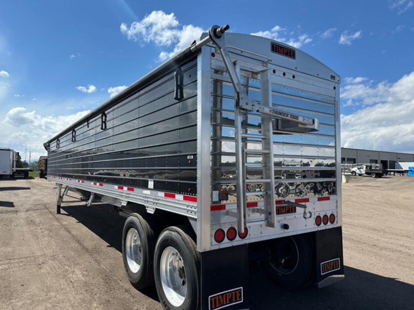 Trailer rear and side view showing Timpte hopper trailer with polished metal panels, rear ladder, taillights, and badges; visible text includes TIMPT E at top center, Timpte on bumper, license plate area with reflective tape.