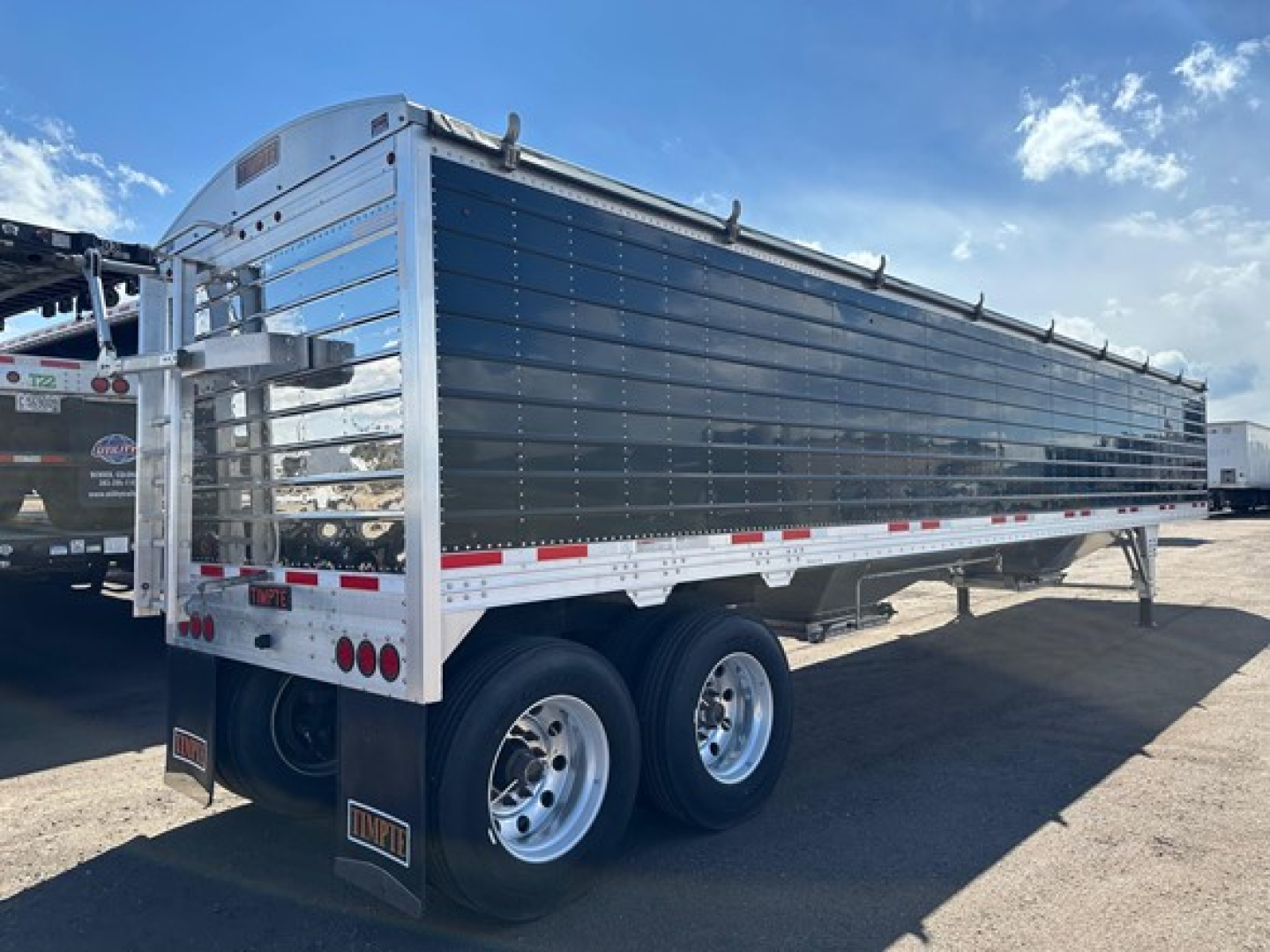 Trailer side view showing a 2024 Timpte Super Hopper W dual-axle aluminum trailer with reflective red/white stripes and a metal ladder/handrail at front.