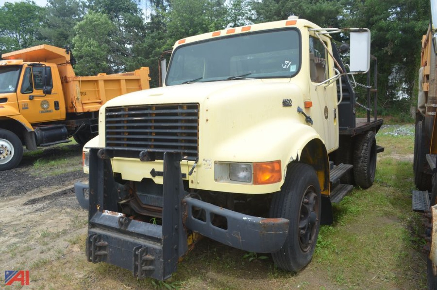 Front view of a cream-colored International 4600 rack truck with a heavy-duty black front bumper and grill, dual rear wheels, and stake bed in a yard.