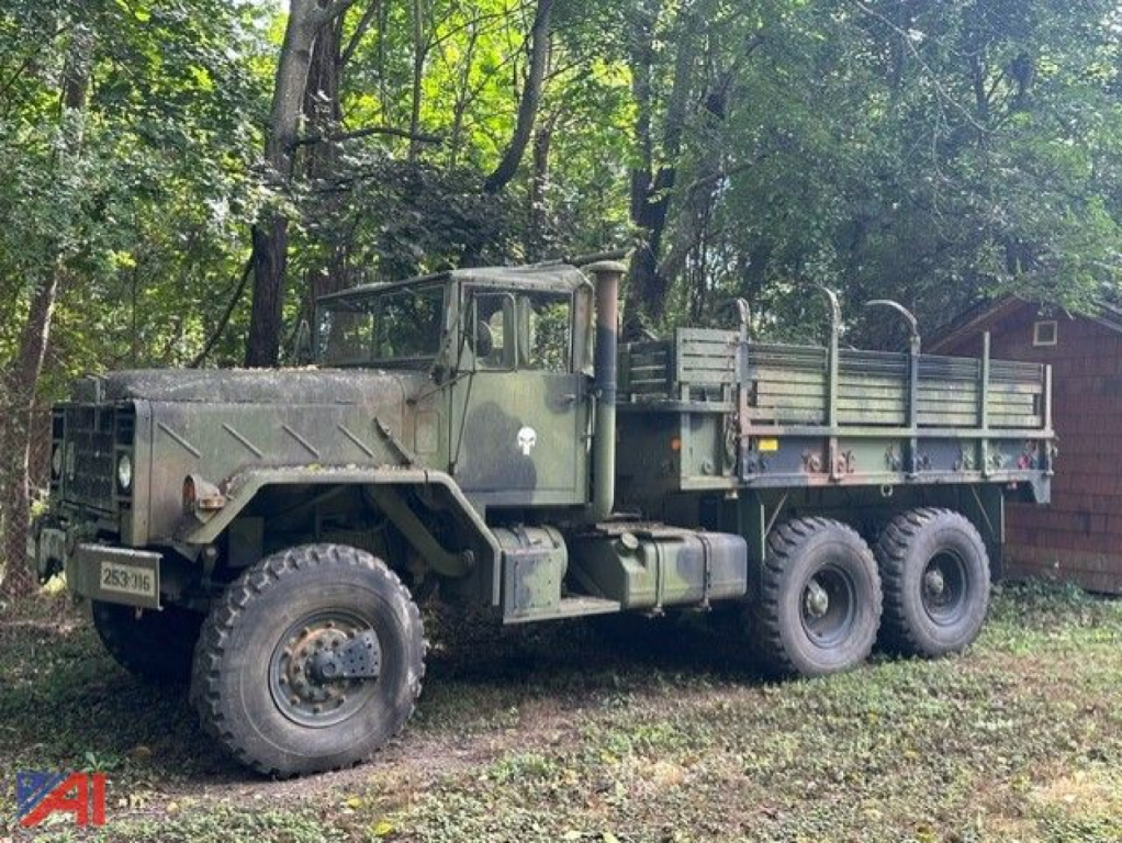 M923A2 or M923AZ cargo truck from Army surplus, olive drab, 6x6 cargo bed with triple rear axles, parked in grass with trees in background.