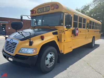 Commercial yellow school bus front-left view with stop sign extended, 'SCHOOL BUS' sign on top, bus number 375, logo, and black bumper. Close-up of front grille and headlight area.