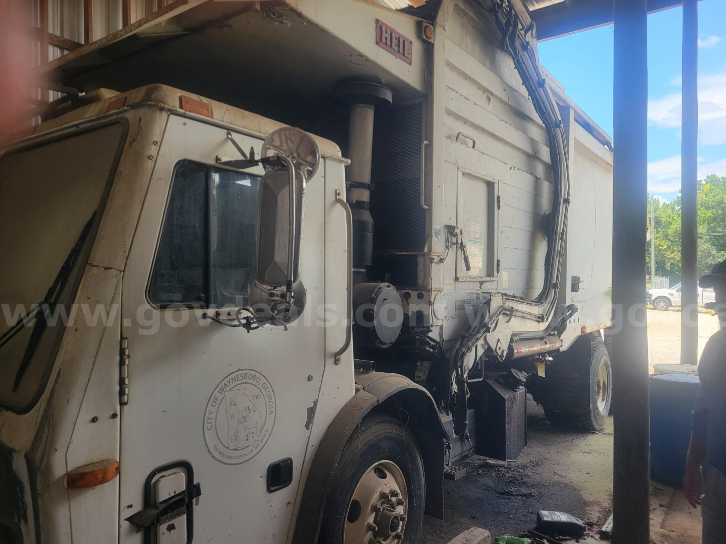 Truck cab and body of a Mack LE613 garbage truck showing side view with hydraulic compactor; visible text includes 'HELI' on top area and circular badge on door.